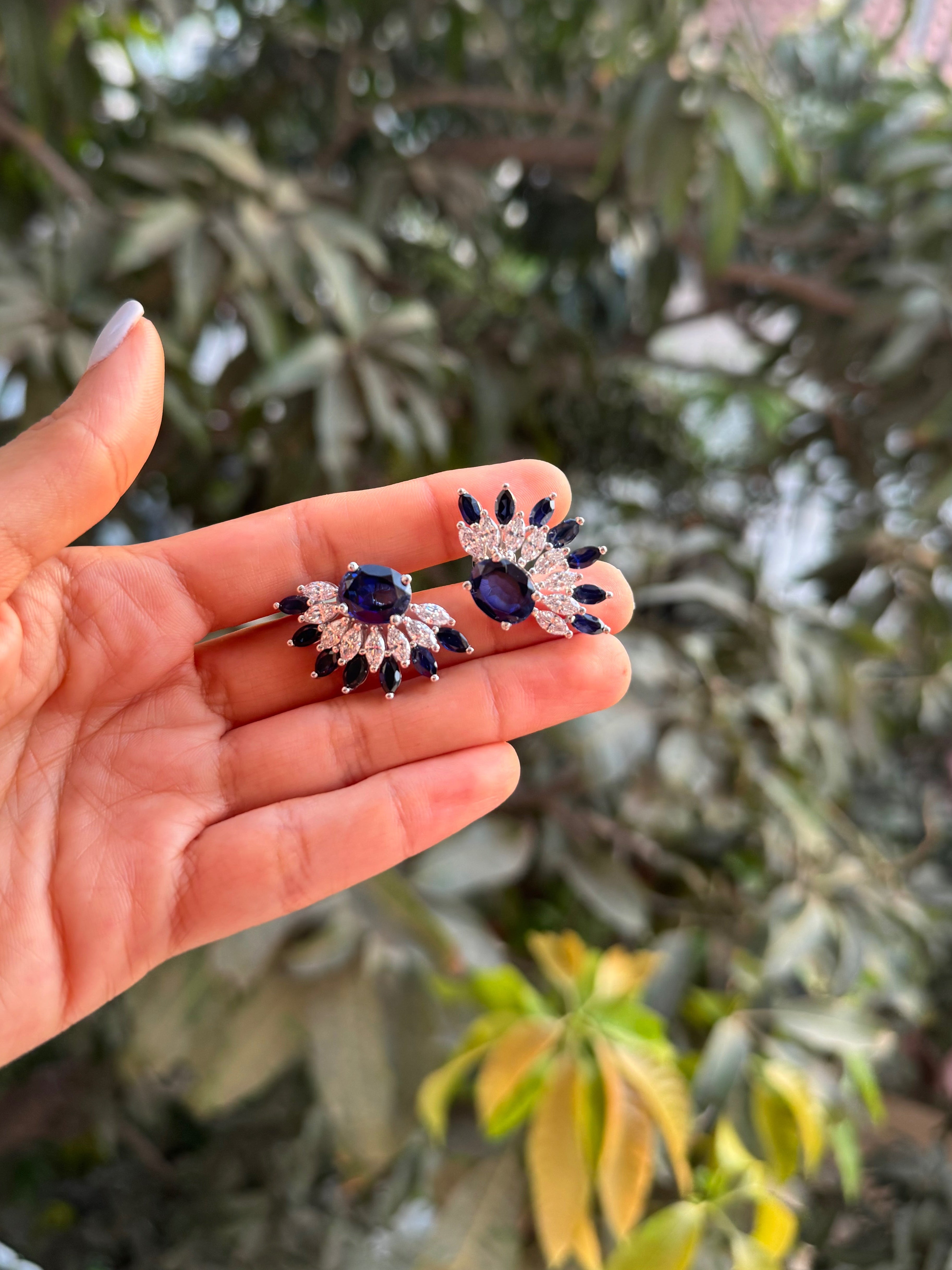 Hand holding a pair of earrings with blue stones against a blurred green leafy background